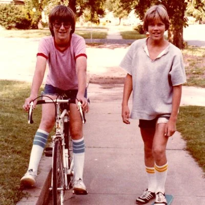Two boys in 1970, one on a bike, the other on a skateboard.